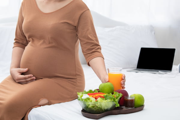 Eine schwangere Frau sitzt mit gesunden Essen und einen Laptop auf dem Sofa.