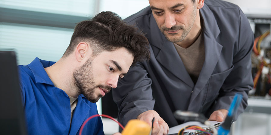 Auszubildender arbeitet an elektronischem Bauteil, ein Ausbilder gibt Hinweise