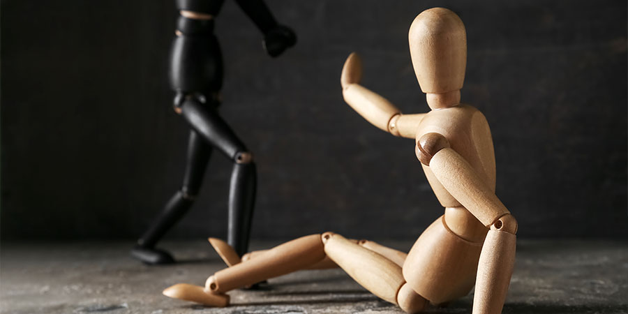 Eine schwarze Holzfigur greift eine am Boden sitzende, helle Holzfigur an