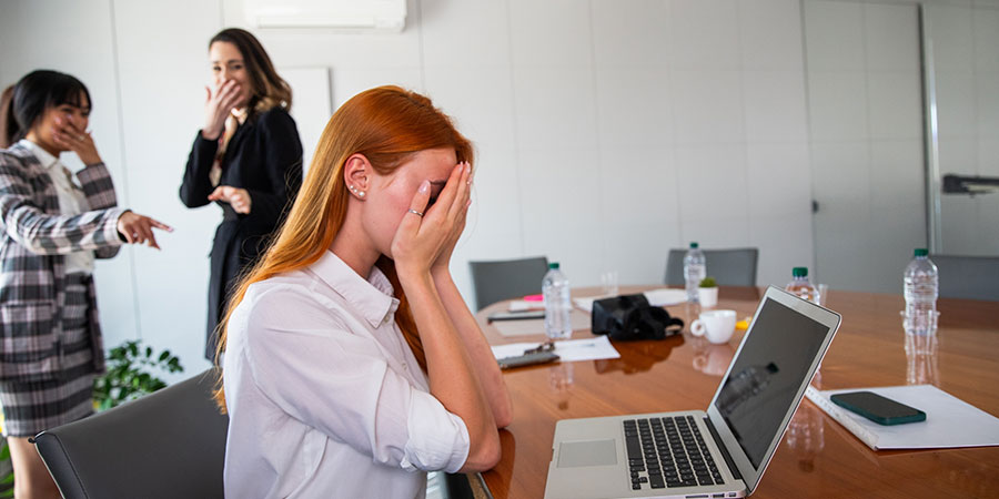 Frau am Arbeitsplatz hält Hände vor das Gesicht und wird von zwei Kolleginnen ausgelacht