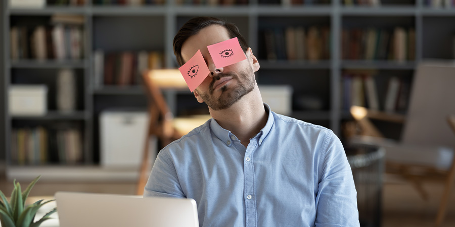 Müder Mann vor Labtop mit Post-Its im Gesicht die Augen darstellen.